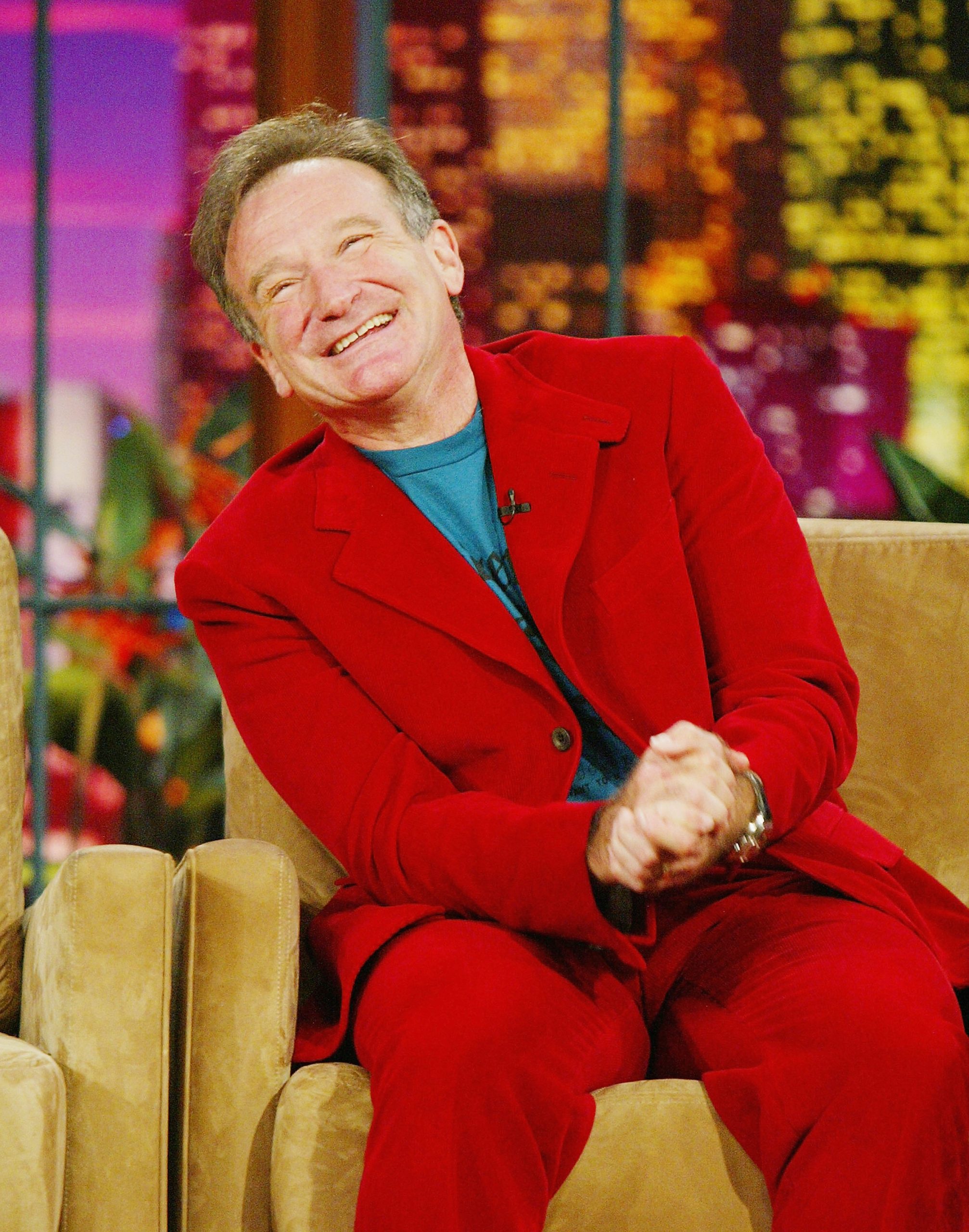 Comedian Robin Williams in a bright red suit sits laughing on a mustard yellow sofa on the set of a late night talk show.