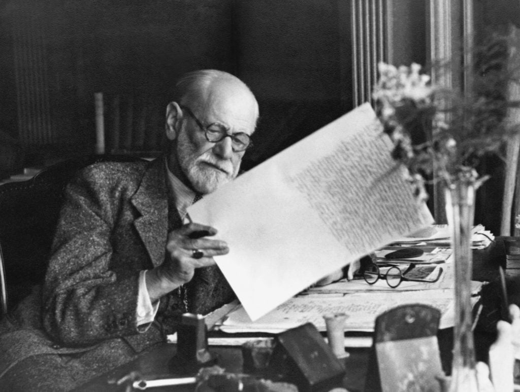 Sigmund Freud sits in a room holding a piece of paper which he is reading, he has glasses on and a desk is cluttered with belongings