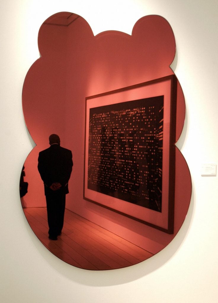 a reflective red material hanging on a white wall, you can see a person and a painting in the reflection, the outline of the object somewhat resembles the abstracted outline of a hippo