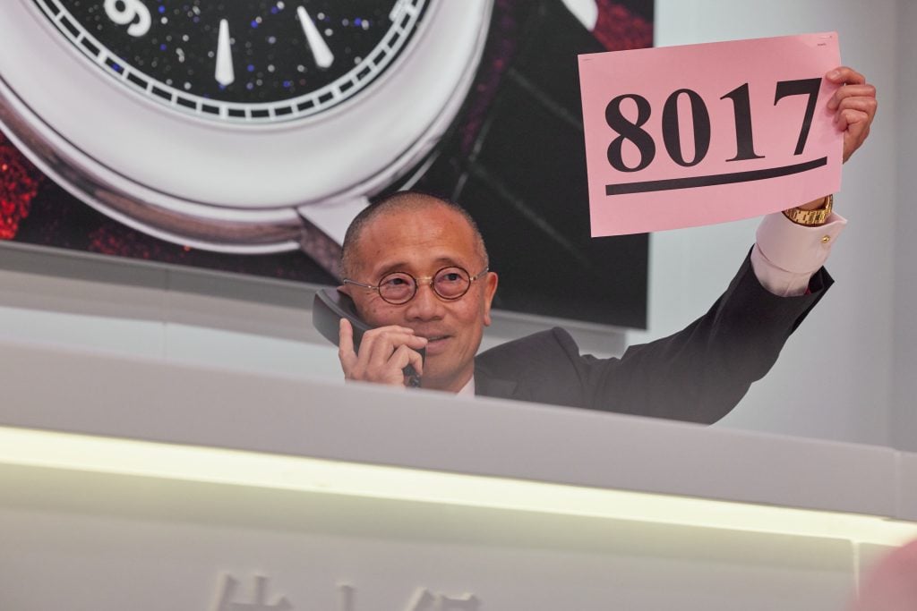 An East Asian man in glasses on the phone on one hand and holding a piece of pink paper that says 8017 with another.