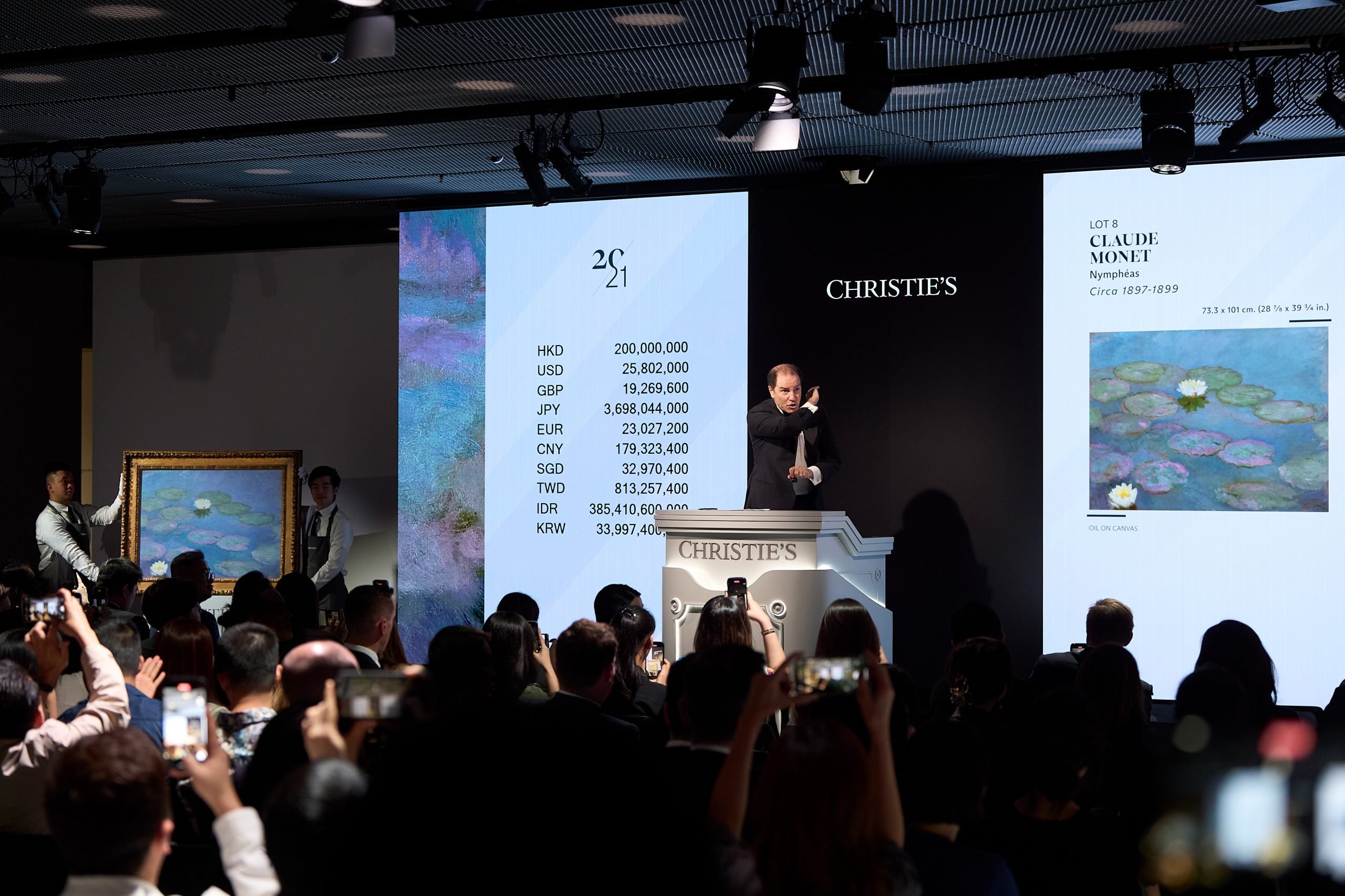 A caucasian man holding a gavel up at a Christie's auction of a Monet waterlilies painting, with the crowd held their smart phones up snapping the moment.