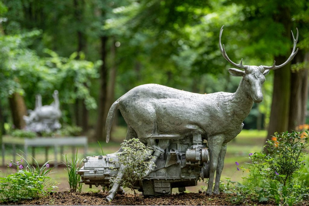 a metal sculpture of a deer with a dense mechanical underbelly is installed in a luscious green forest-like park