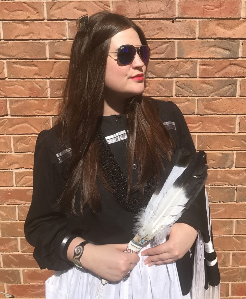 Raven Halfmoon has long brown hair in sunglasses holding two feathers and stands against a brick wall