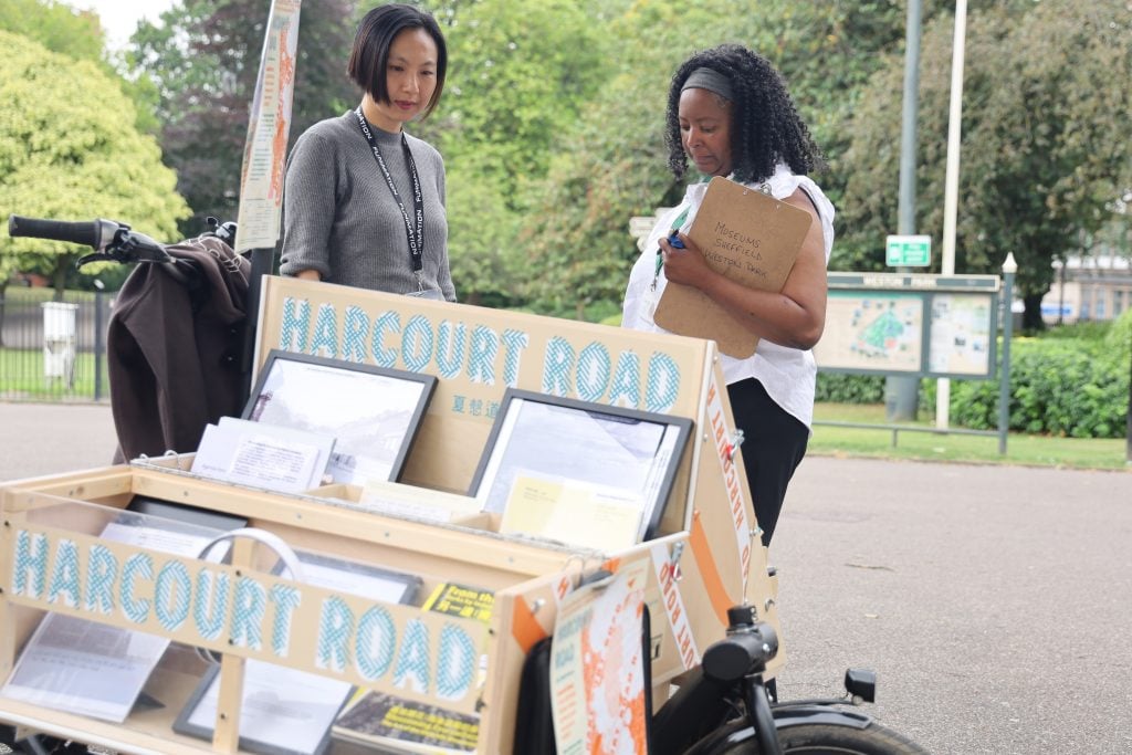 A short hair East Asian woman standing next to a woman of African descent, in front of them is a cart that spells Harcourt Road