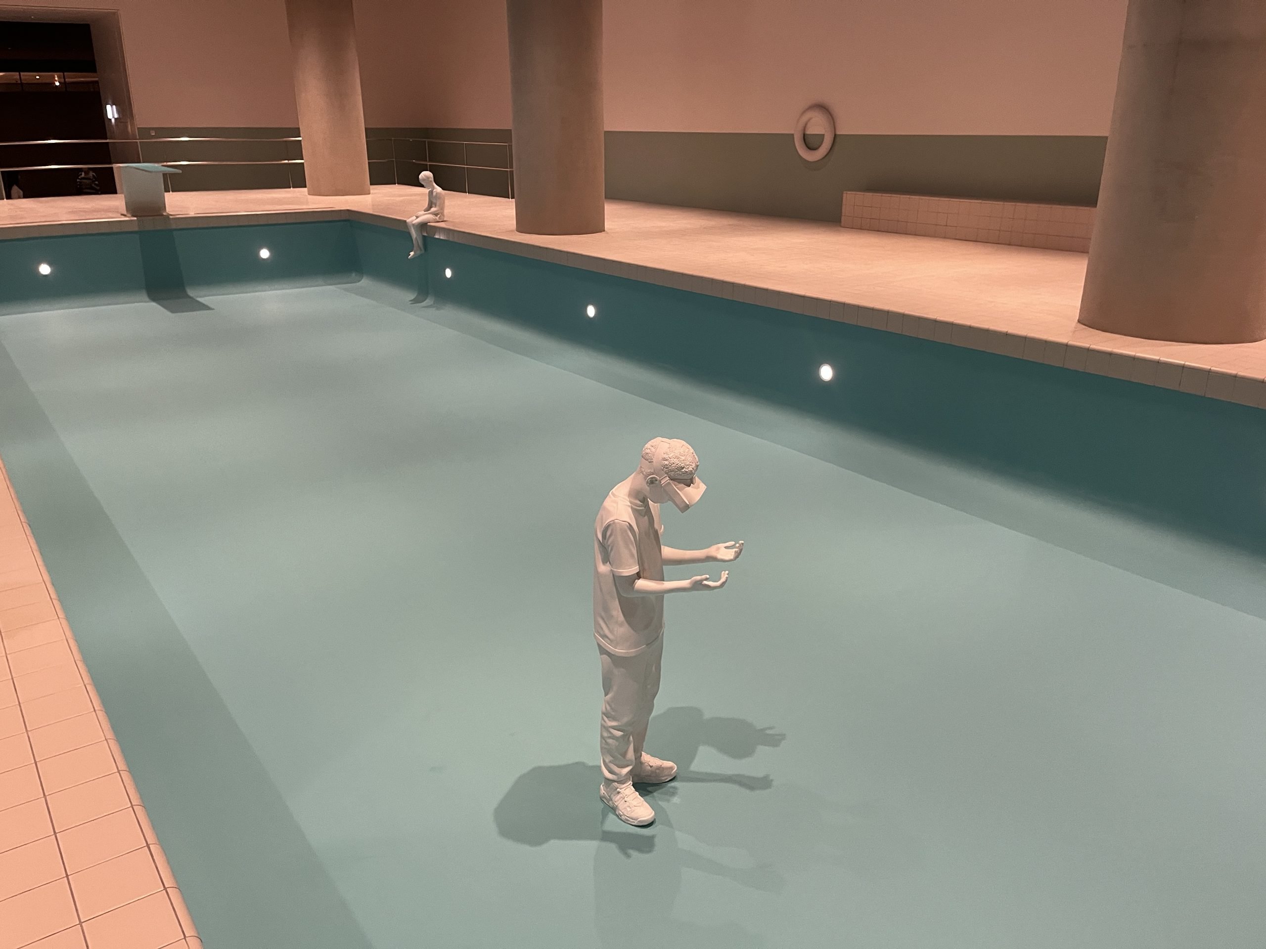 Two gray-white sculptures of boys reside in an empty indoor pool with blue floors and walls.