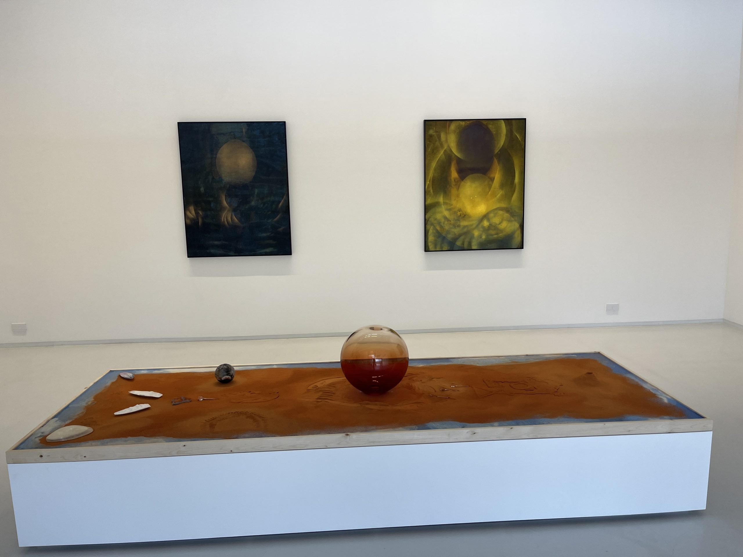 Two abstract, celestial paintings hang on a white wall in front of a white plinth with a glass sphere