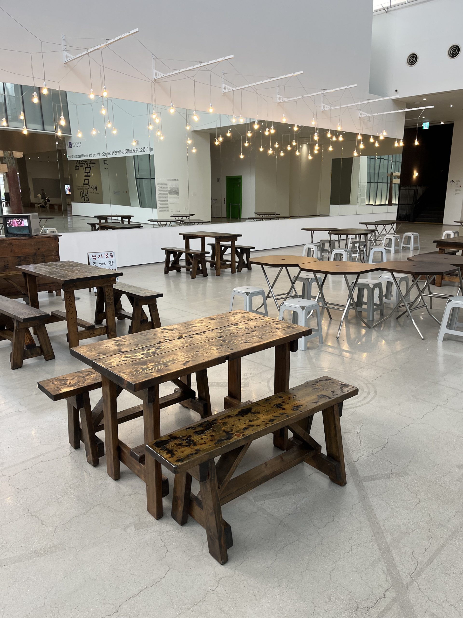 Rustic wooden tables and chairs sit in a white-walled gallery next to mirrors and strings of lights.