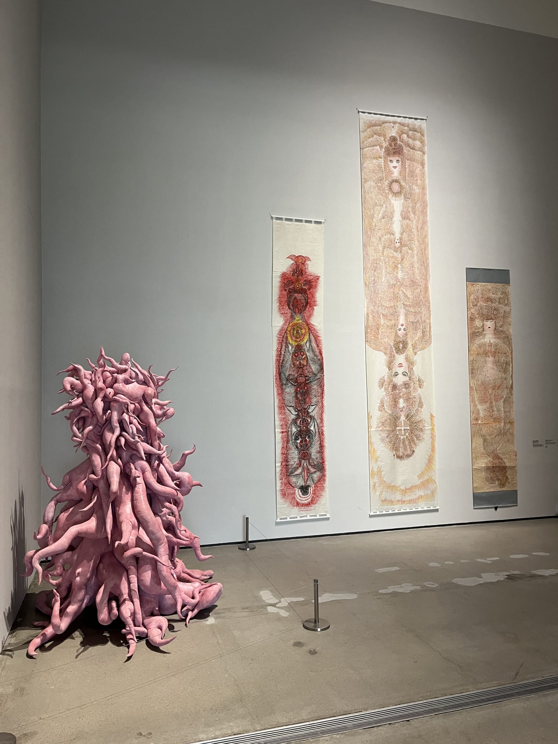 A pink sculpture of many tentacles sits on the floor next to drawings with mysterious, frenetic beings in a gallery with high ceilings