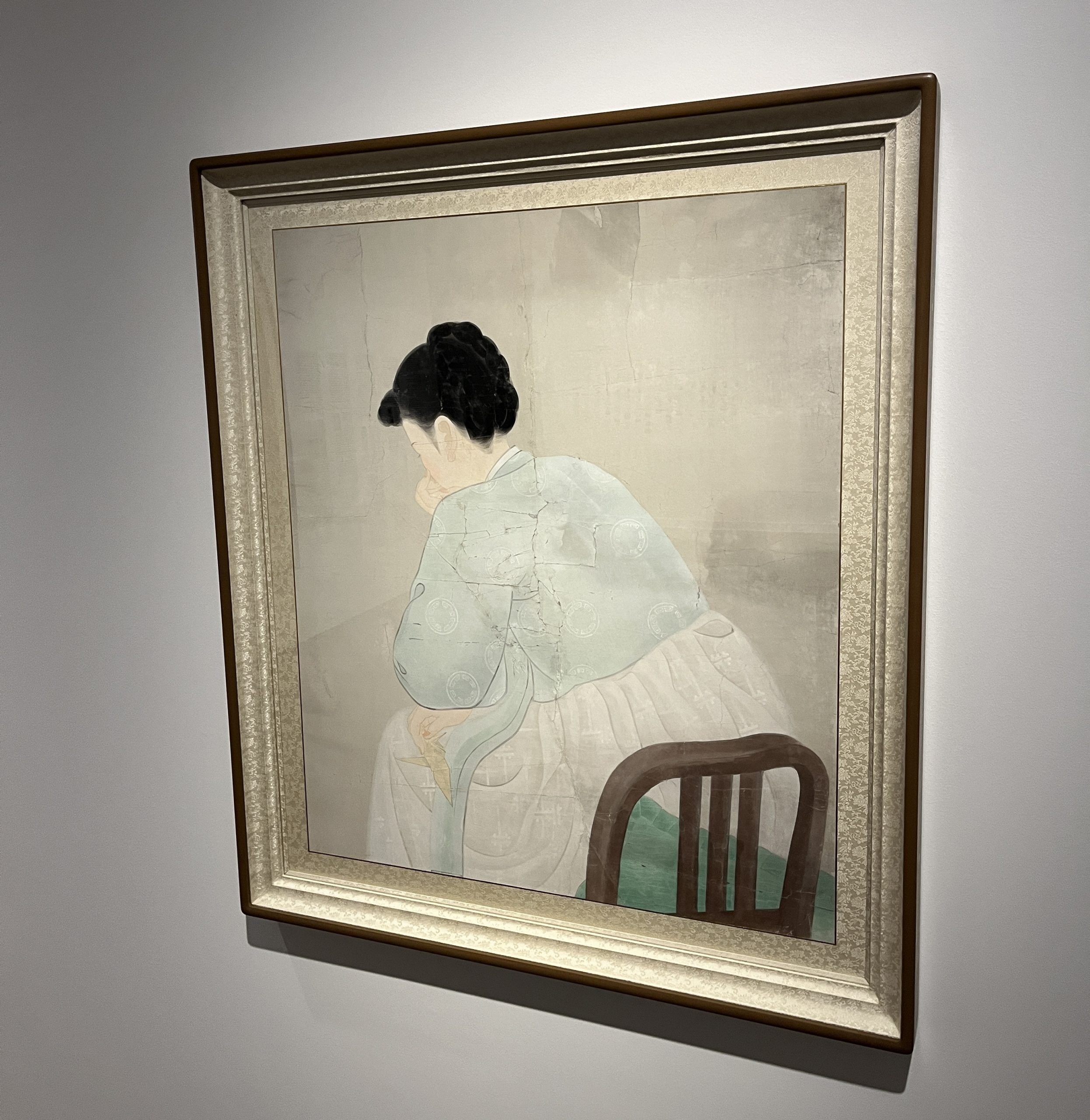 A woman in a hanbok with a blue top sits on a chair, leaning over, looking away in a painting.