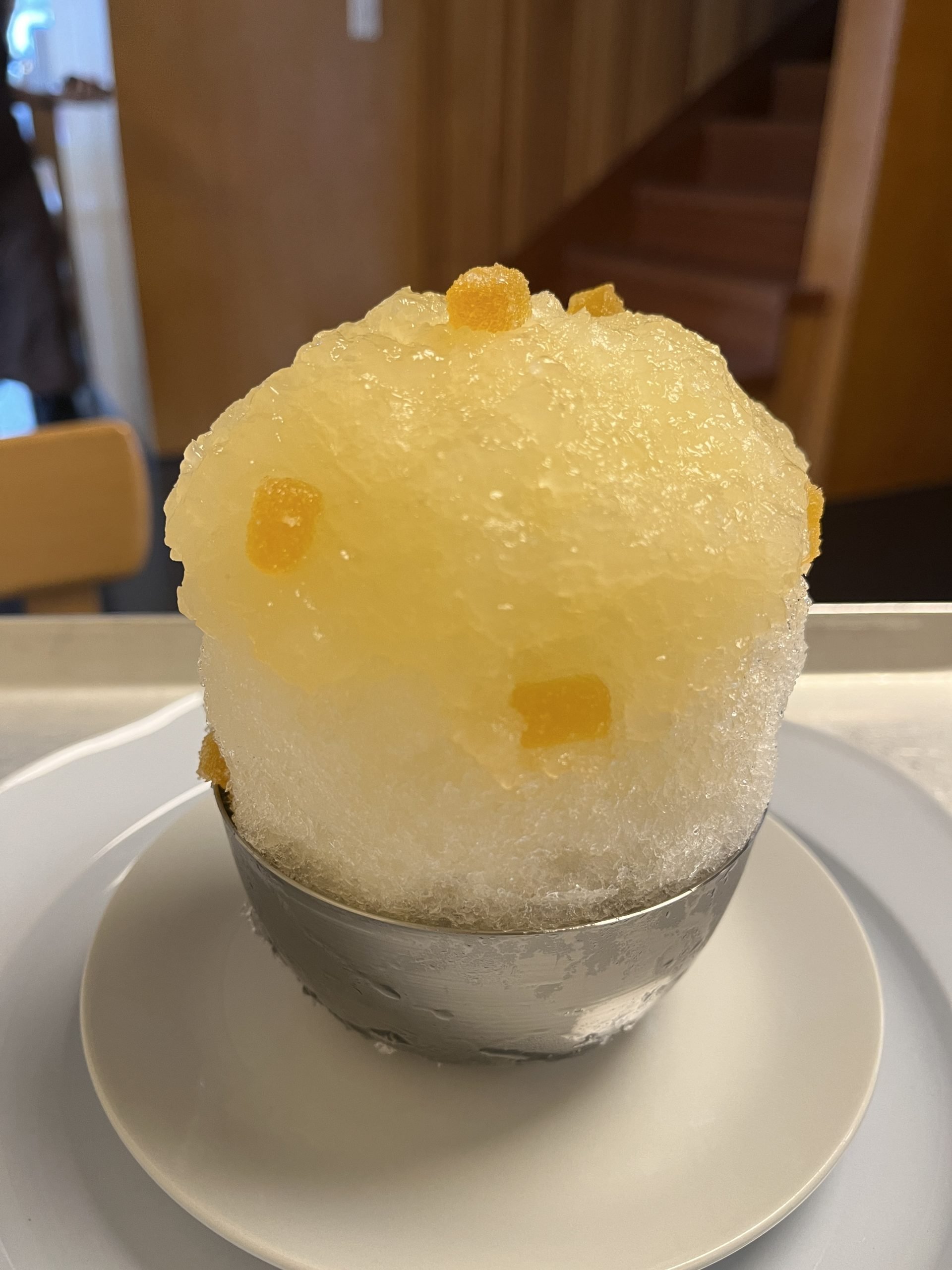 A massive stack of shaved ice sits in a metal bowl with a yellow color atop it and small yellow candy lozenges.