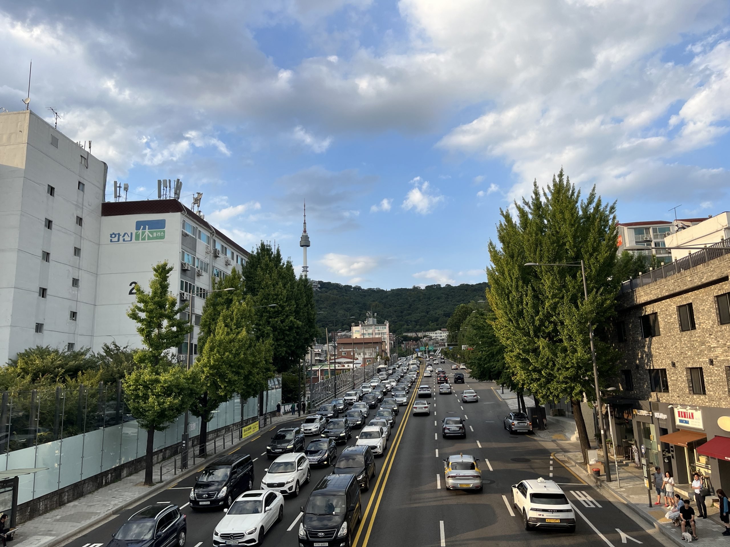 A cityscape shots a blue sky with clouds, a tower atop a hill and cars and buildings up close