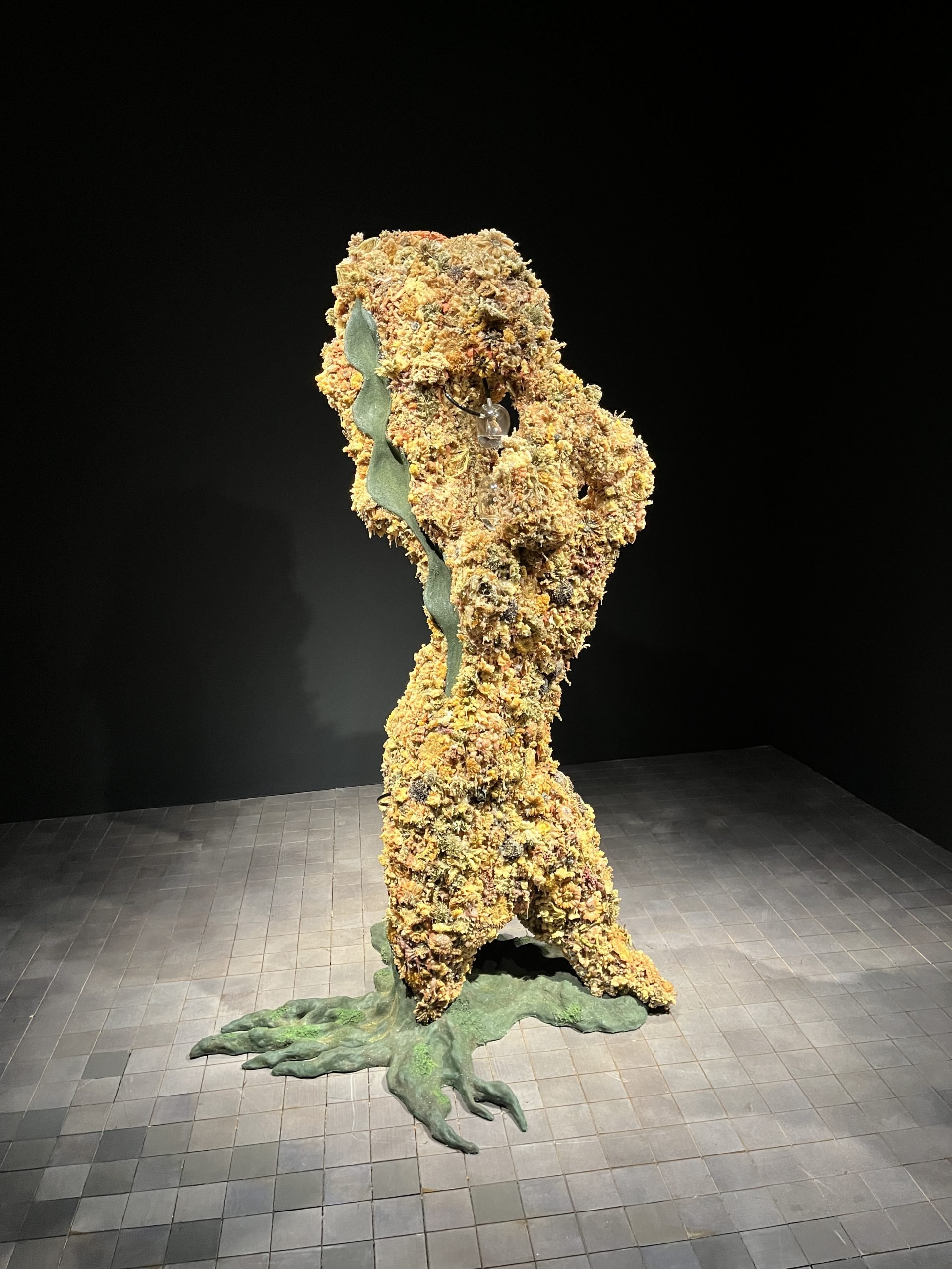 A mysterious standing shape made of green and yellow flowers is spotlit in a dark room.