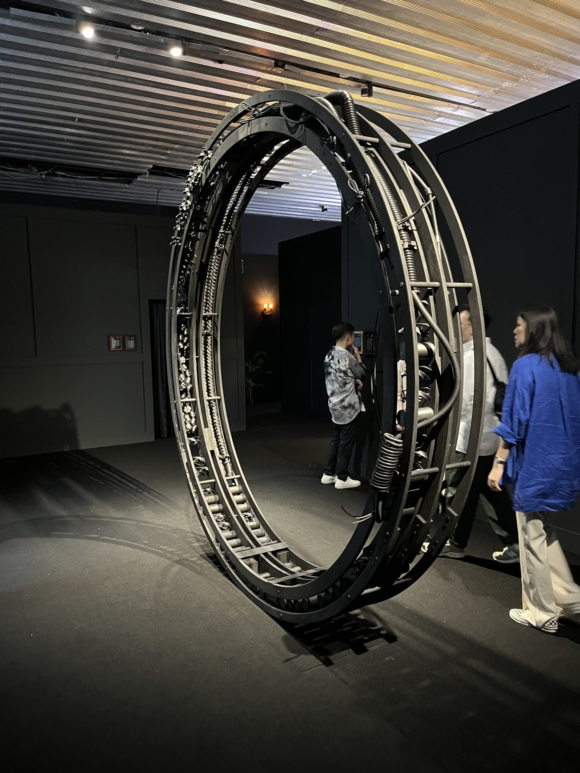 A dark gallery holds a large ring-like sculpture that is standing up. People stand around it.