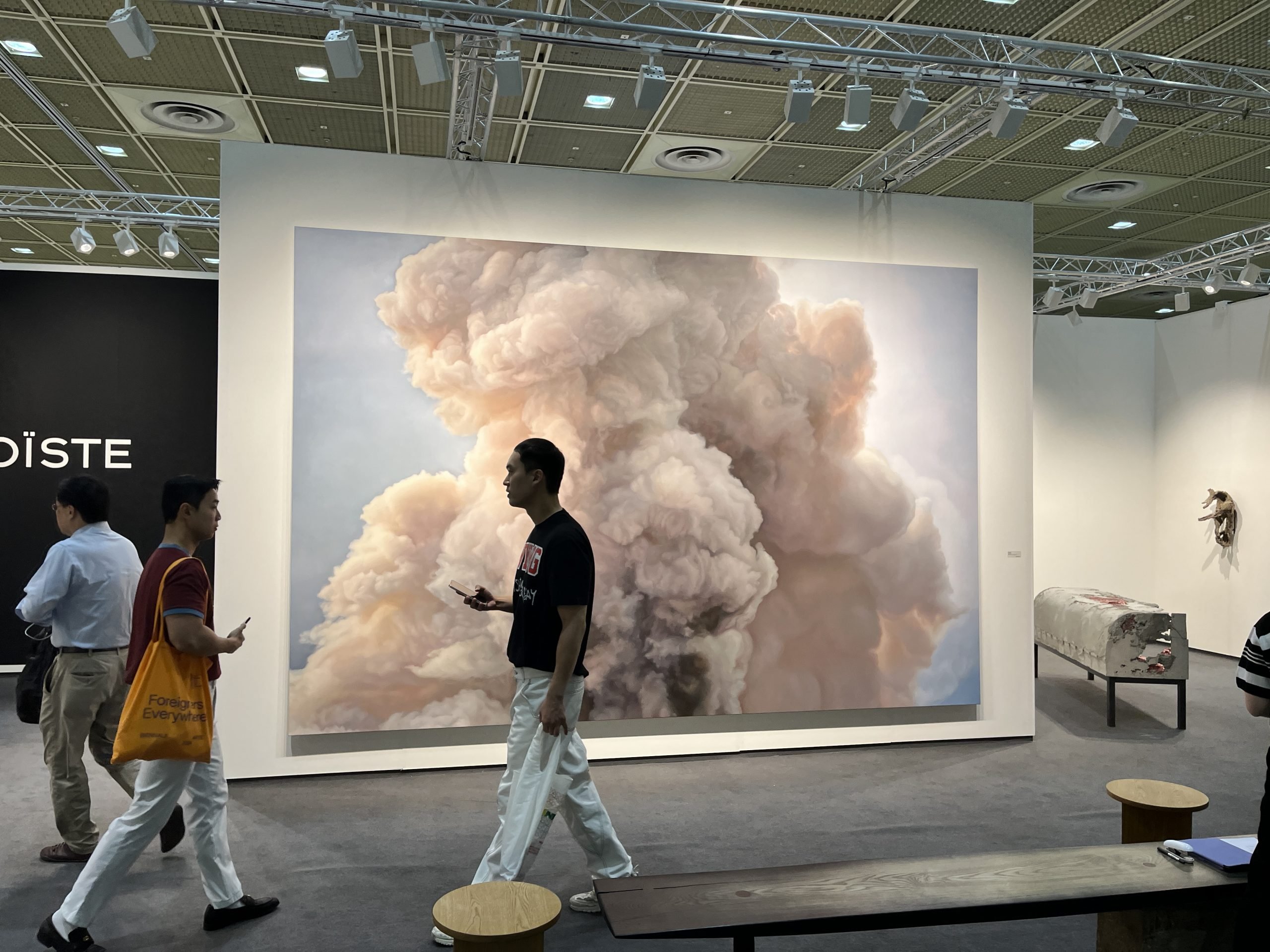 A massive convention center holds a white-walled art fair booth with a large painting of clouds or smoke