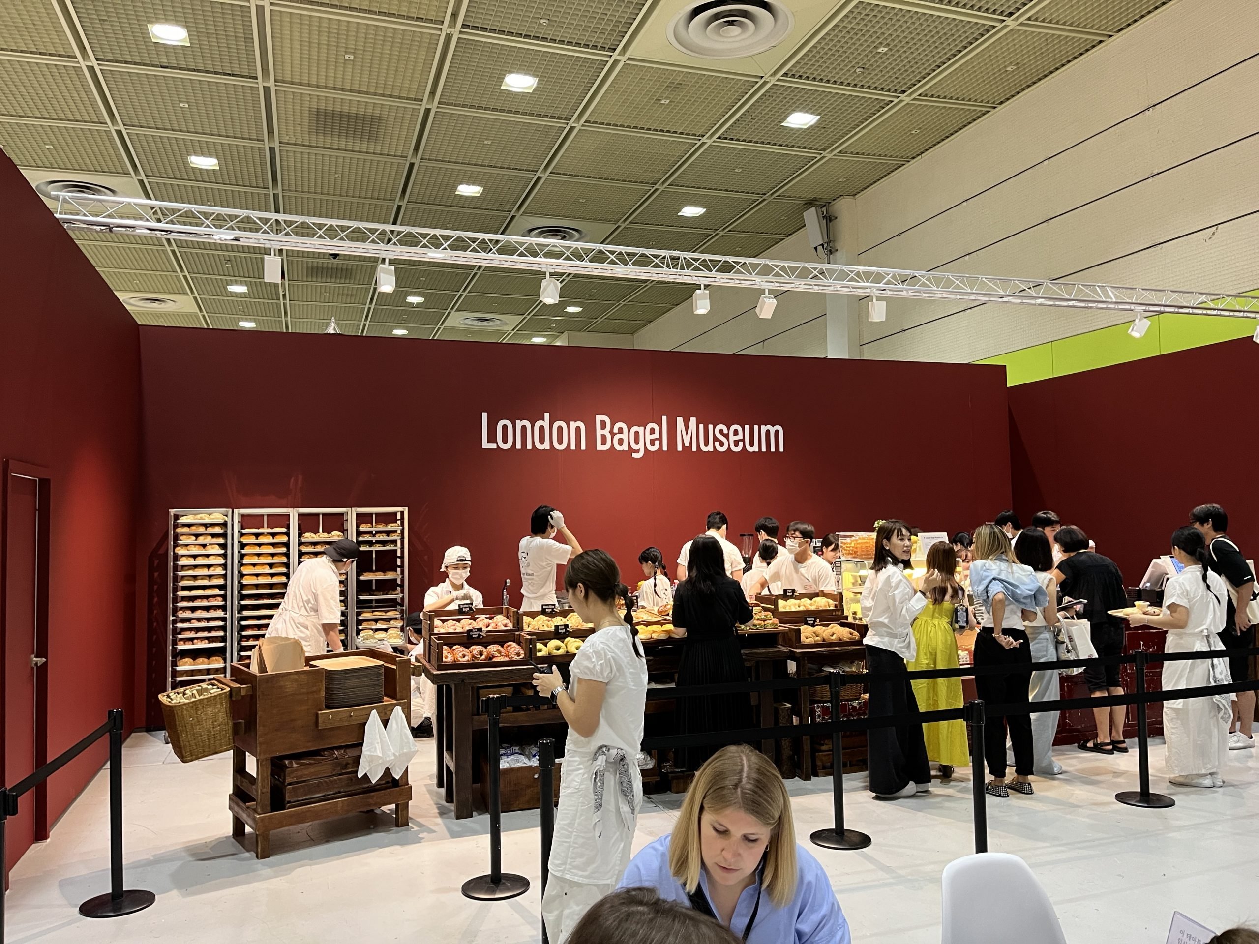 A burgundy wall has white text that reads "London Bagel Museum." People wait in line to procure bagels.