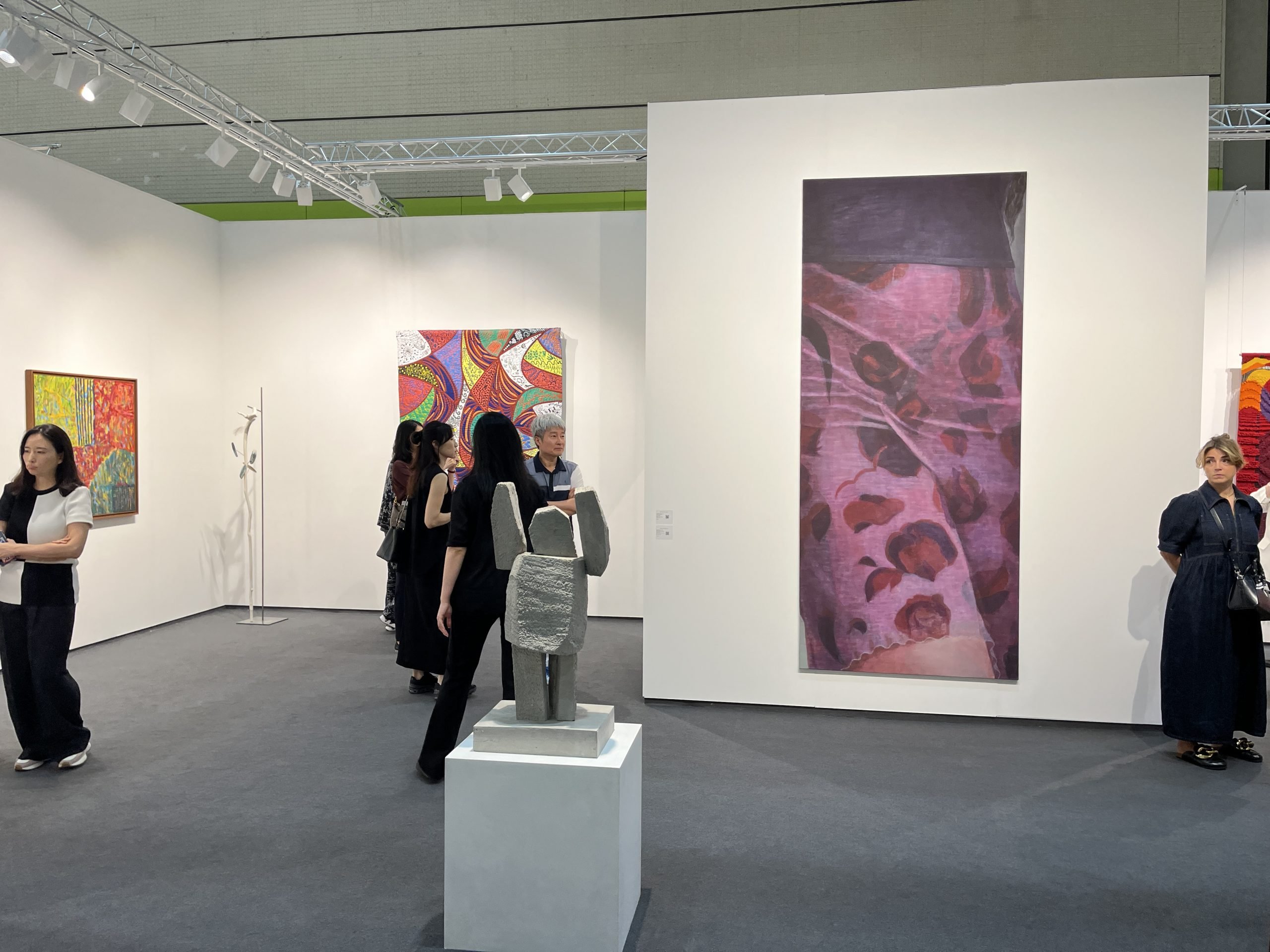 A white-walled art fair booth holds paintings and a small gray sculpture