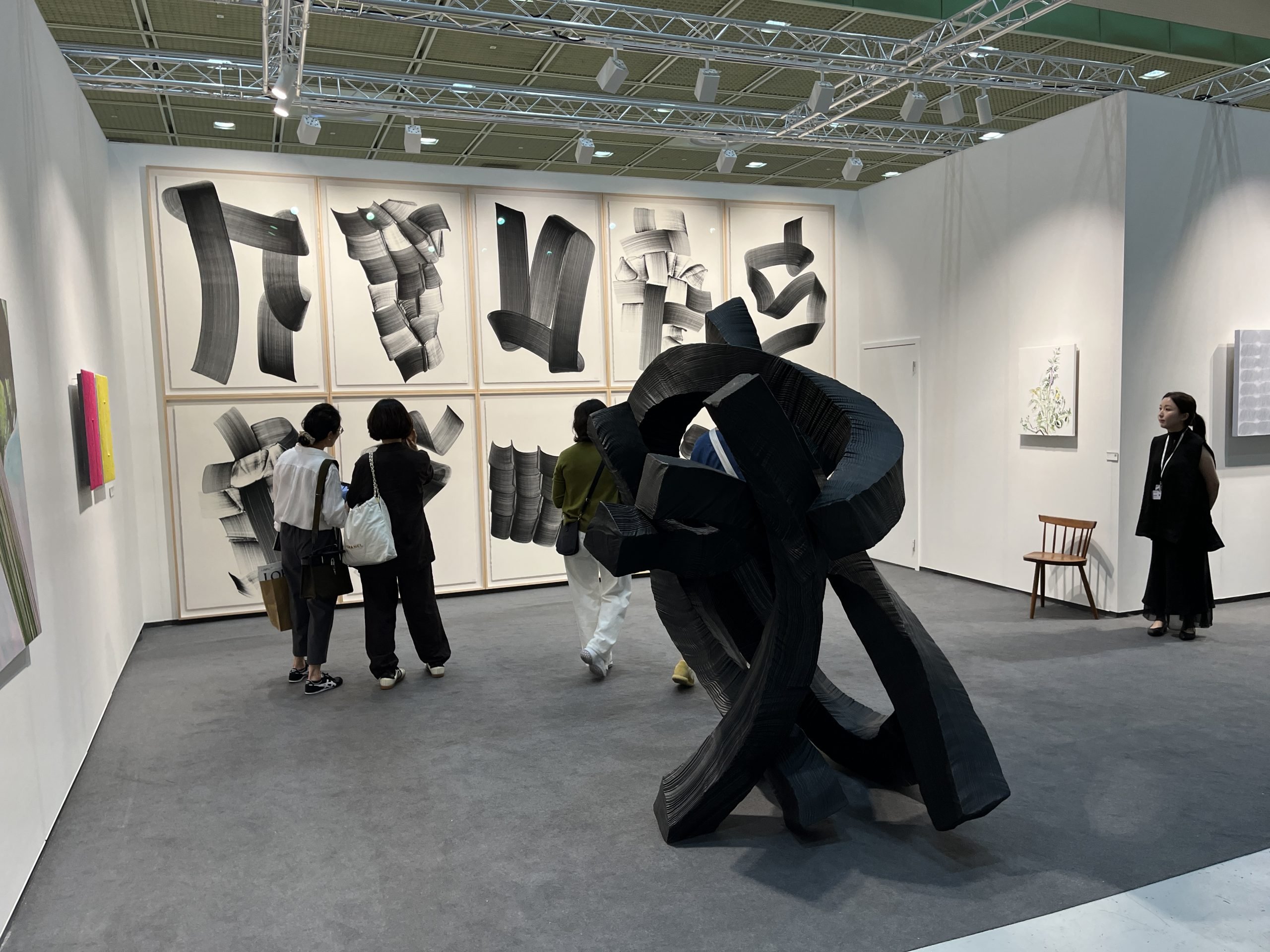 A tangled, black, abstract sculptures sits in an art fair booth in front of a white wall holding a number of framed works with similar black forms on white