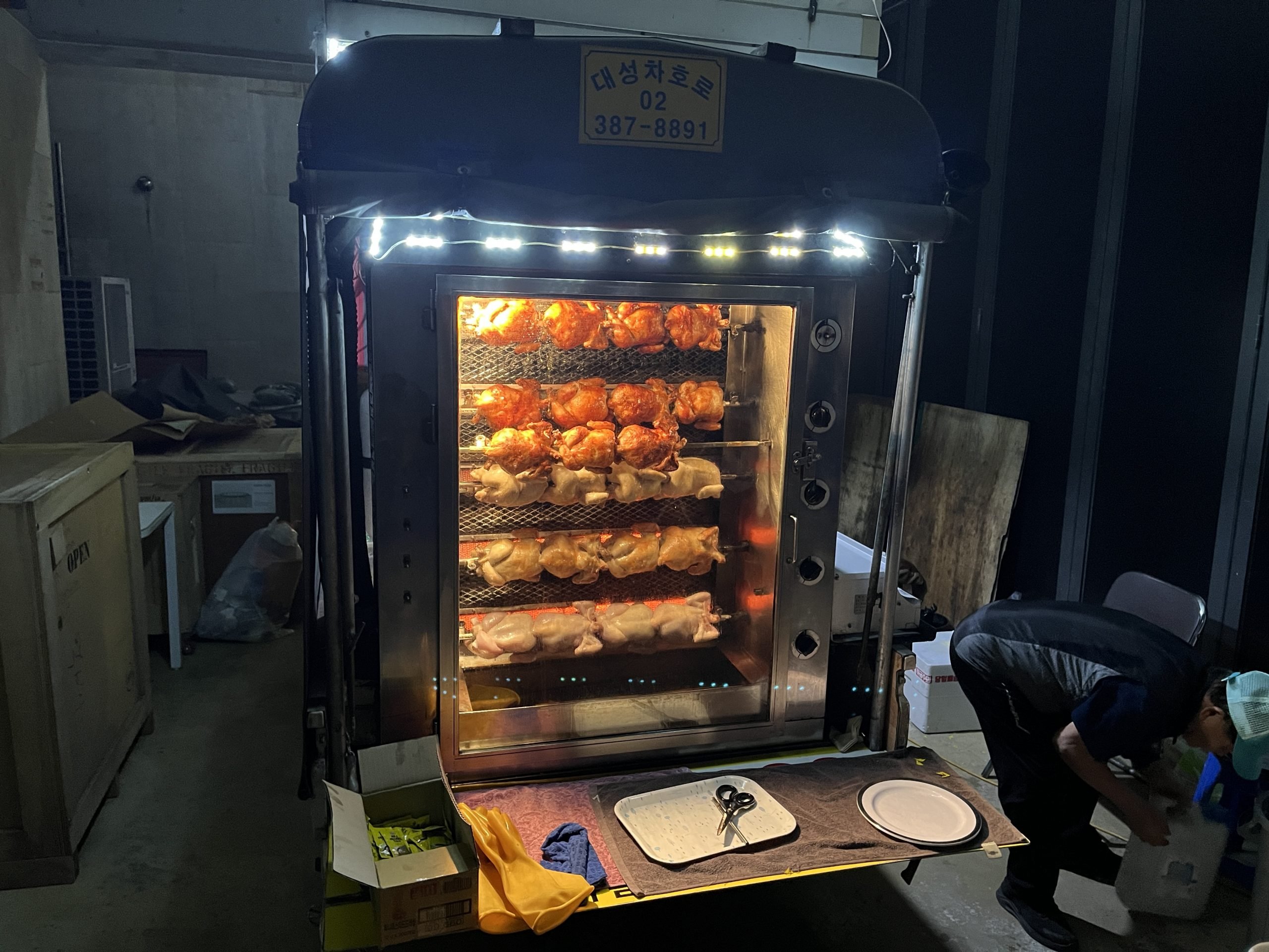 Dozens of chickens roast at the back of a truck on a dark night. A man is at work in the corner.