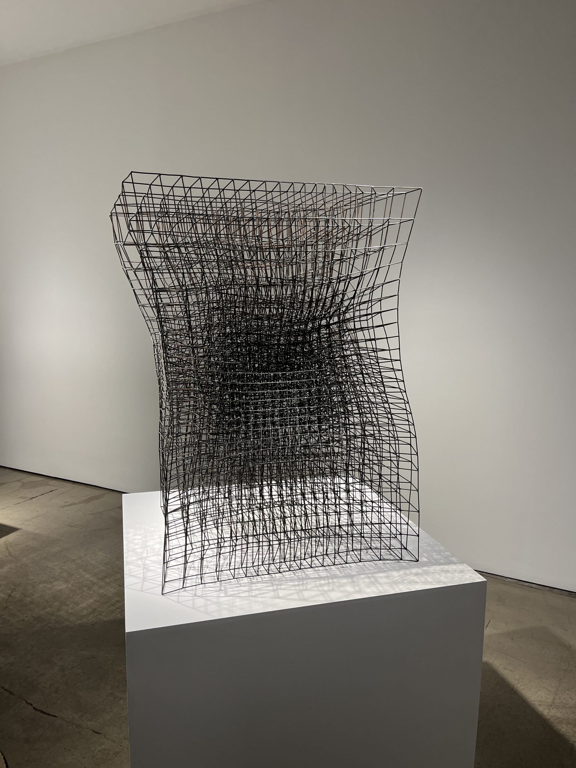 A densely gridded cube of metal wires that is slightly wavy sits on a white plinth.