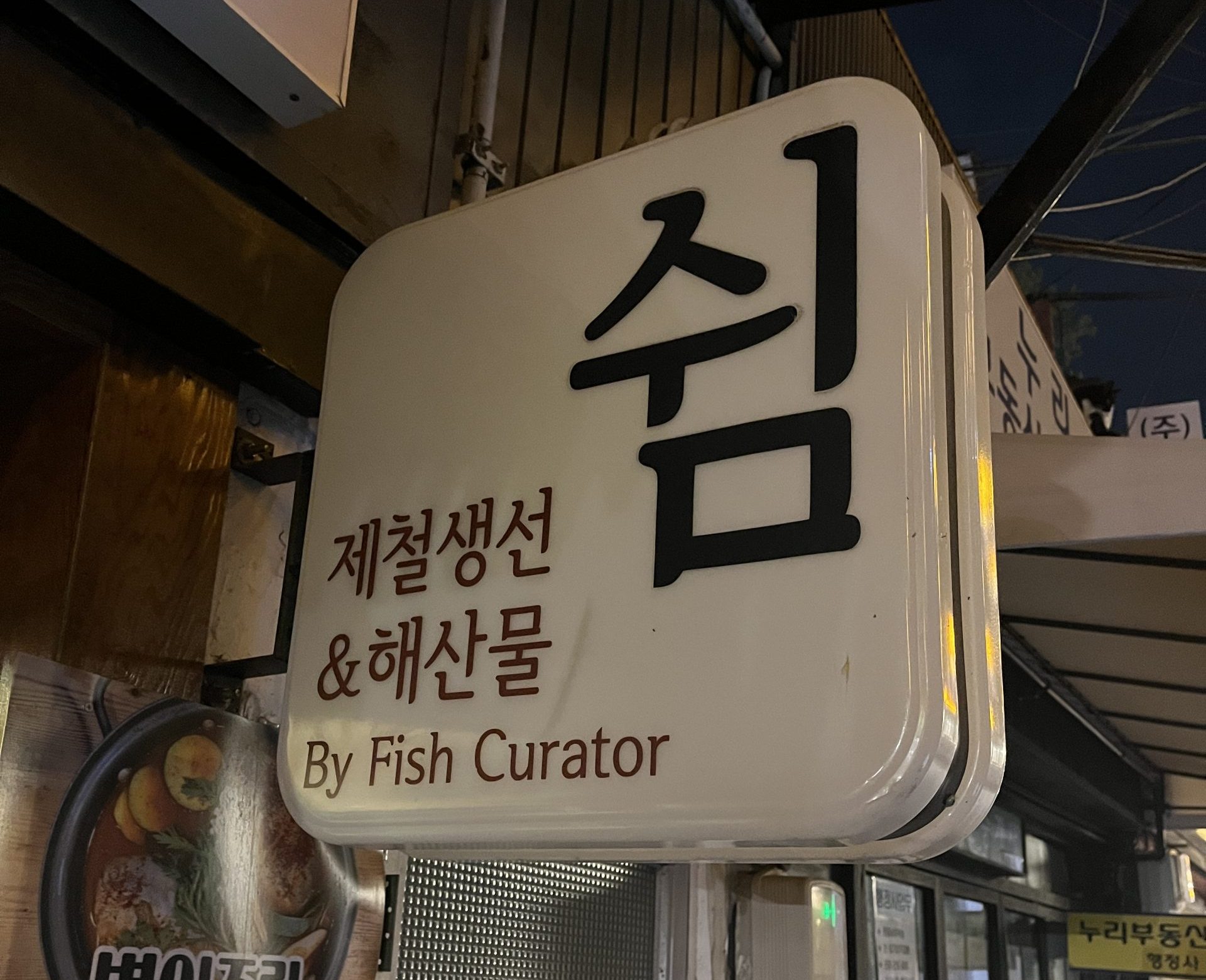 A small white street sign has hangul and three English words: "By Fish Curator"