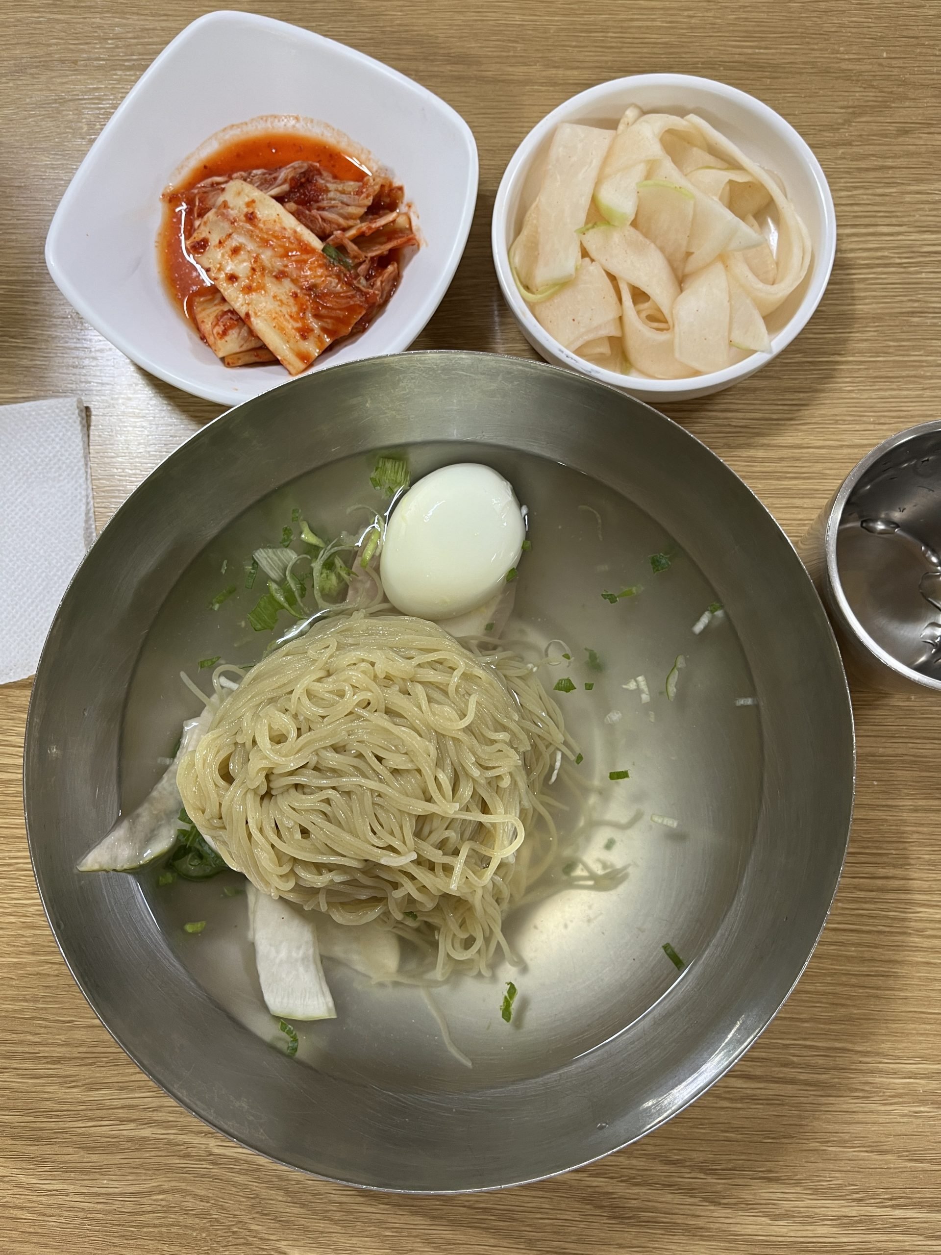 An overhead view of a bowl with clear broth, noodles, and a hard-boiled egg. Two smaller plates hold red and white kimchi.