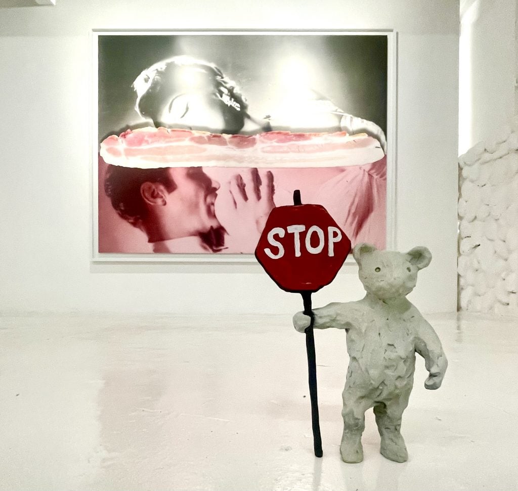 This image shows a white bear sculpture holding a stop sign, standing in front of a large monotone photo at the back hung on a white wall