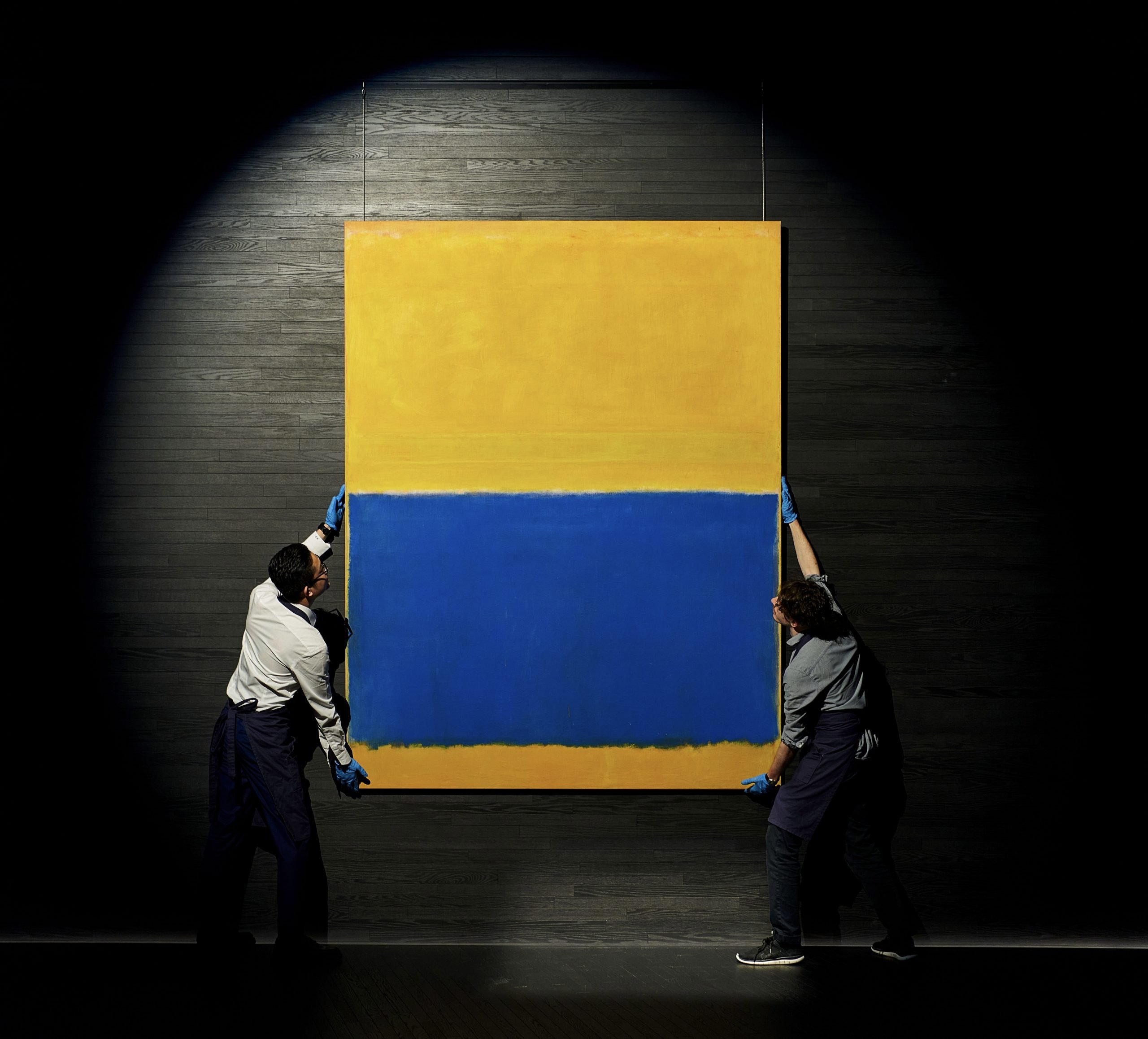 Two men holding a large canvas in yellow and blue, each standing on both sides of the painting.