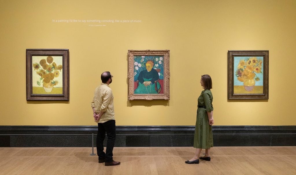 a spacious gallery space with pale yellow walls has two paintings, two are of sunflowers in a vase and the other is a portrait. two visitors to the museum stand and look at the portrait from either side of it