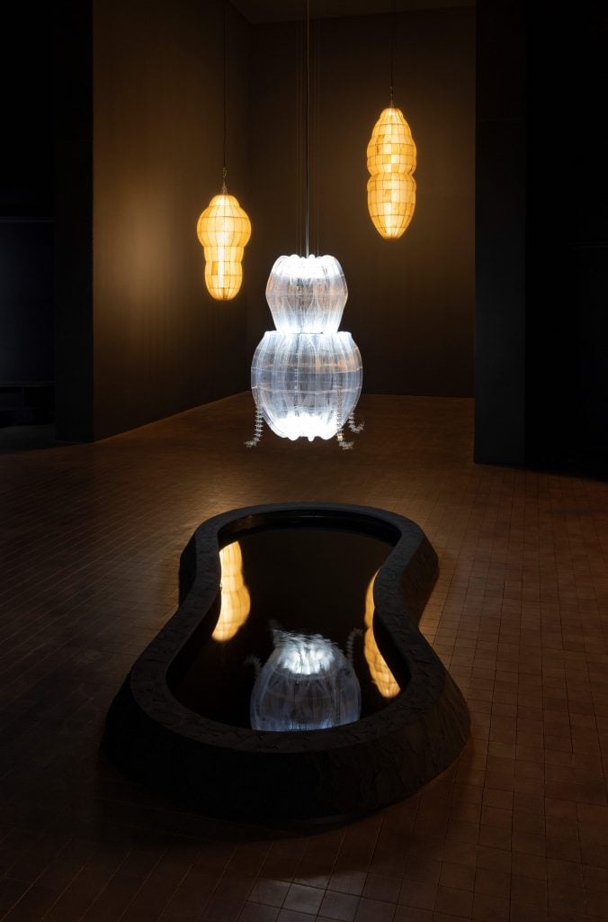 Three installation works that look like lanterns hung from the ceiling in a dark exhibition room, hovering over a pond of water.