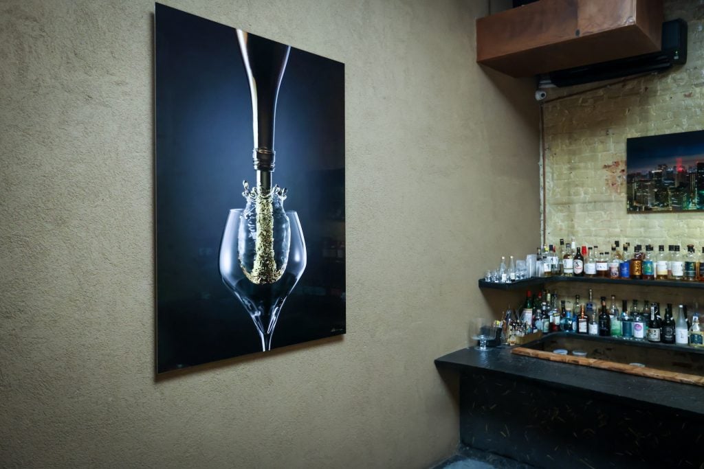 Installation view of "Nathan Myhrvold: Intention and Detail" in the lounge space at the Gallery by Odo, New York. A photo of wine splashing into a glass hangs on a wall next to a bar. 