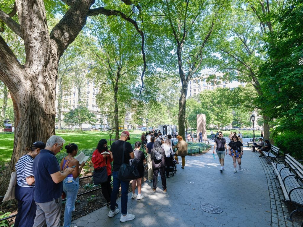 A long line of people waiting on a paved path in the park to take their portrait with JR's "Inside Out Project" photo booth truck for "Portraits on Climate and Health: Dreams We Carry" at the American Museum of Natural History in New York.