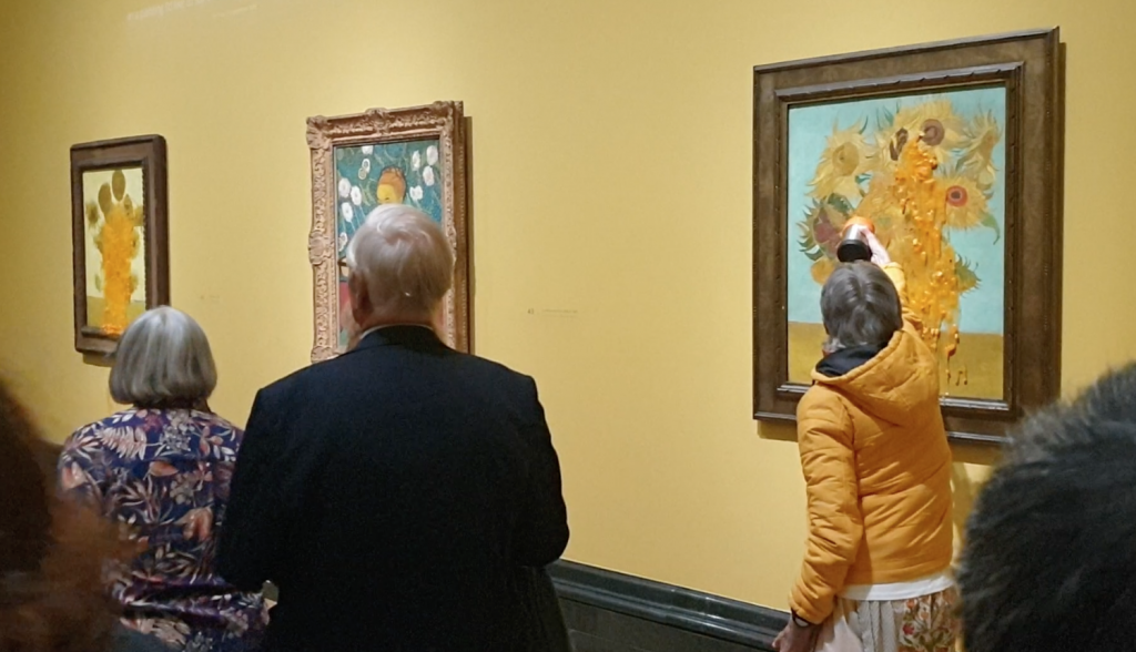 a photograph of a museum interior with paintings hanging on a yellow wall. to the right, a woman is throwing a thick orange liquid at a painting of sunflowers in a vase, there is a small crowd of other people in the room to the left