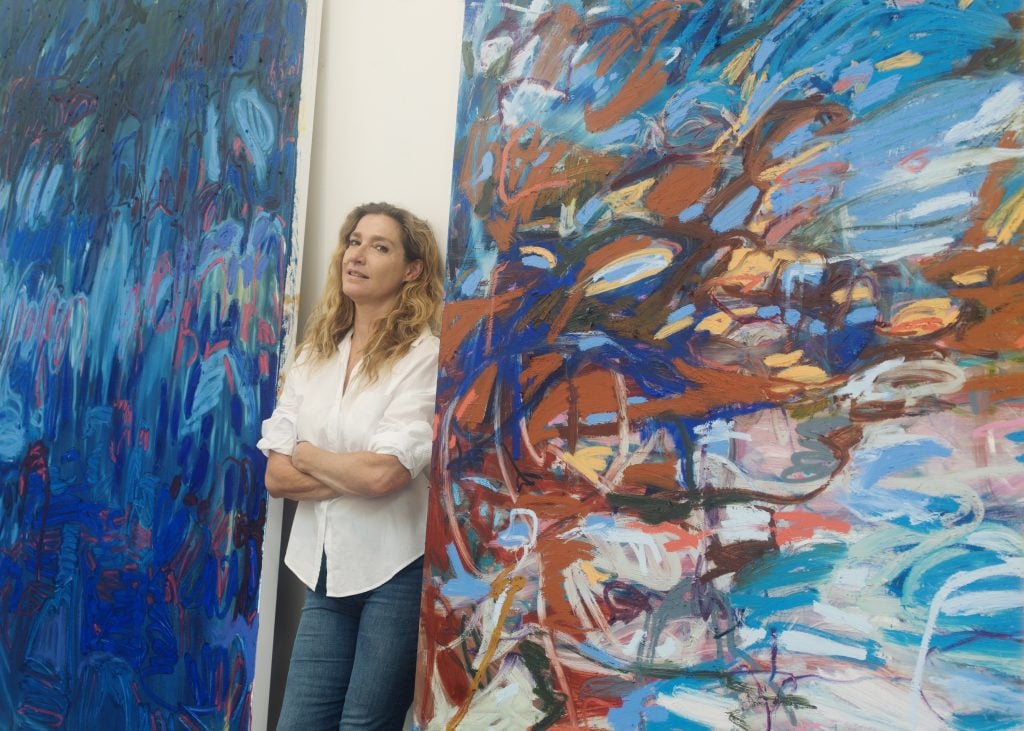 a woman with diry blonde wavy hair stands between two monumental abstract canvases in blues