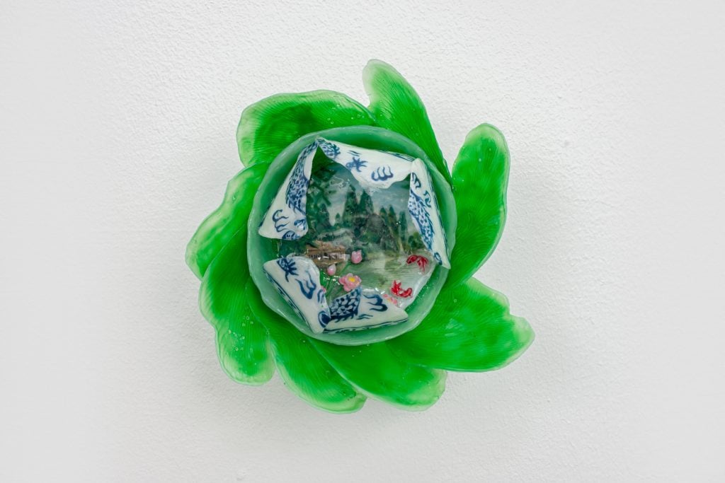 a small shiny object stuck to a wall that has lurid green petal-like forms stick out in a border and colorful layered Chinoiserie blue and white patterns within