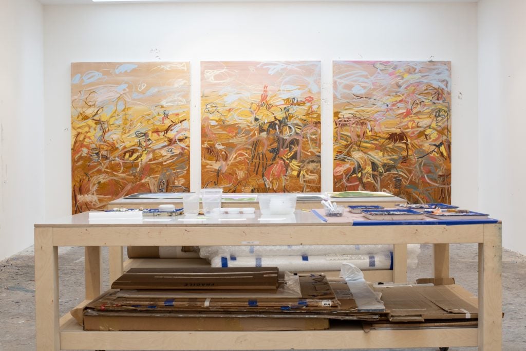 three abstract paintings in yellow, gold, and umber in an artist studio behind a table