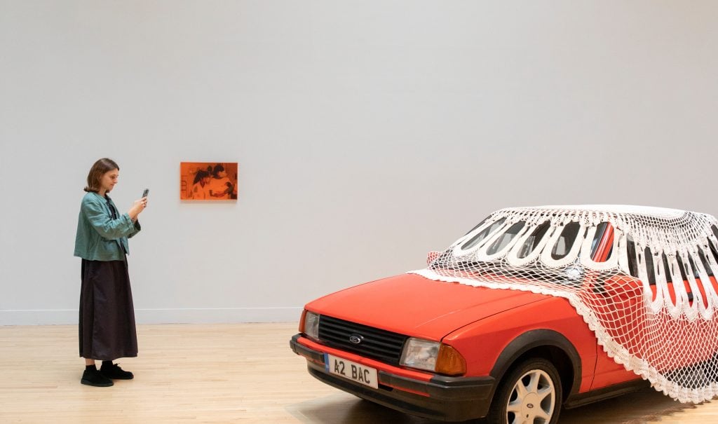 a photograph of a gallery space in which there is a small, vaguely orange image on the far wall and installed in the middle of the room a vintage looking red car with a massive white doilly draped over the top 