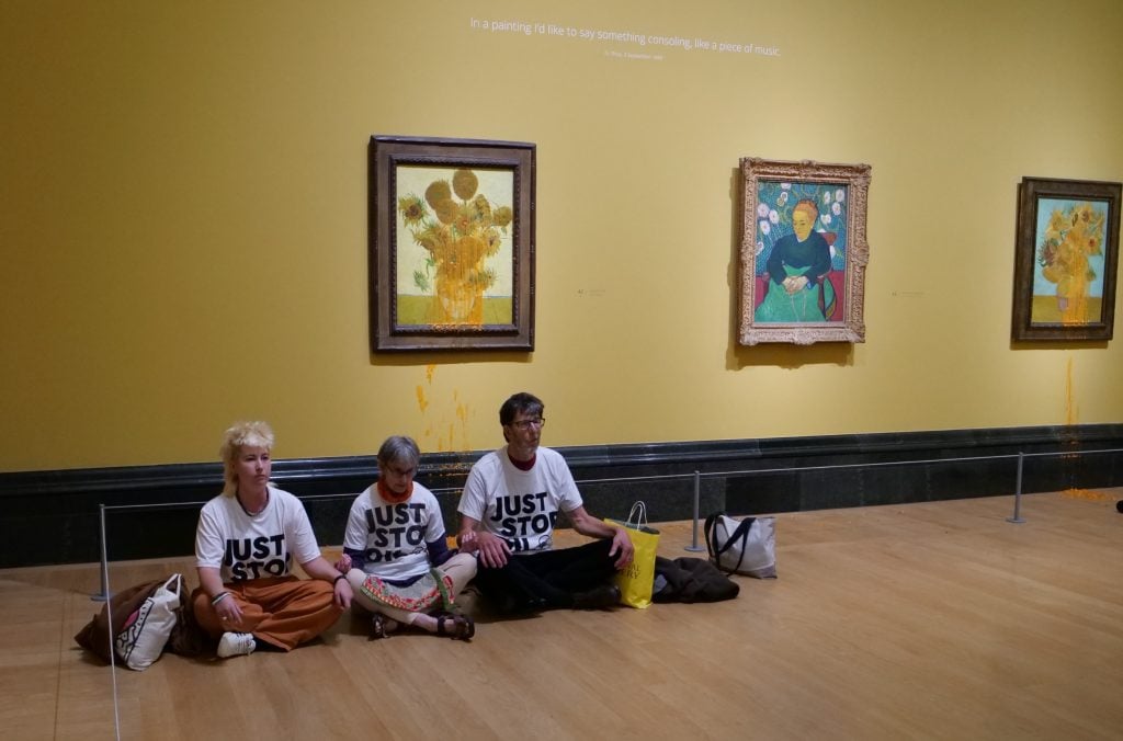 three people wearing white t-shirts reading 'Just Stop Oil' are sitting on the floor of a gallery space in a museum, behind them are three paintings on the wall. Two of the paintings are of sunflowers and have been splattered with a thick orange liquid that is dripping down over the frame and onto the wall beneath