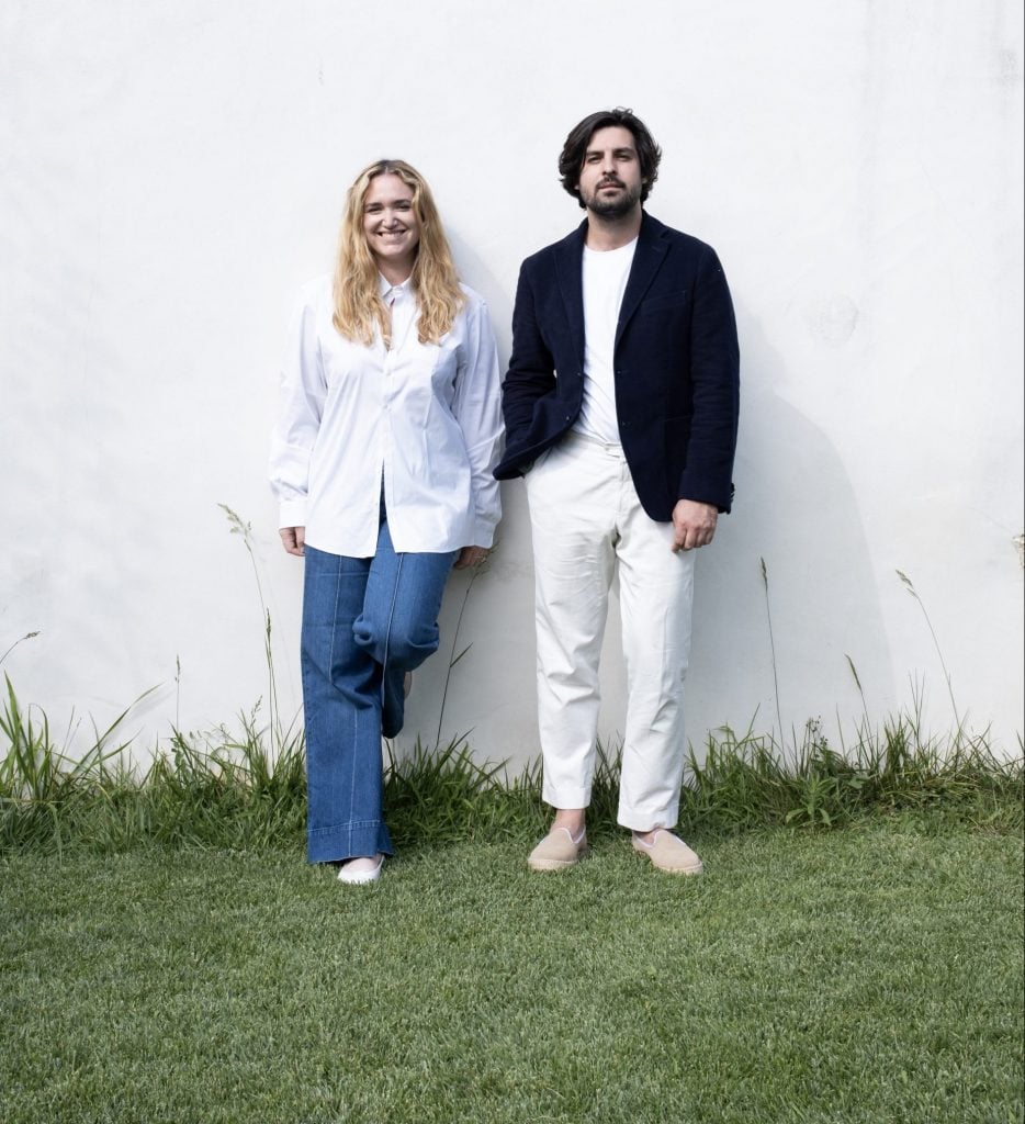 a Oman in jeans and a white blouse with curly blonde hair and a man in a white shirt blue blazer and white trousers are standing in grass leaning against a white wall 