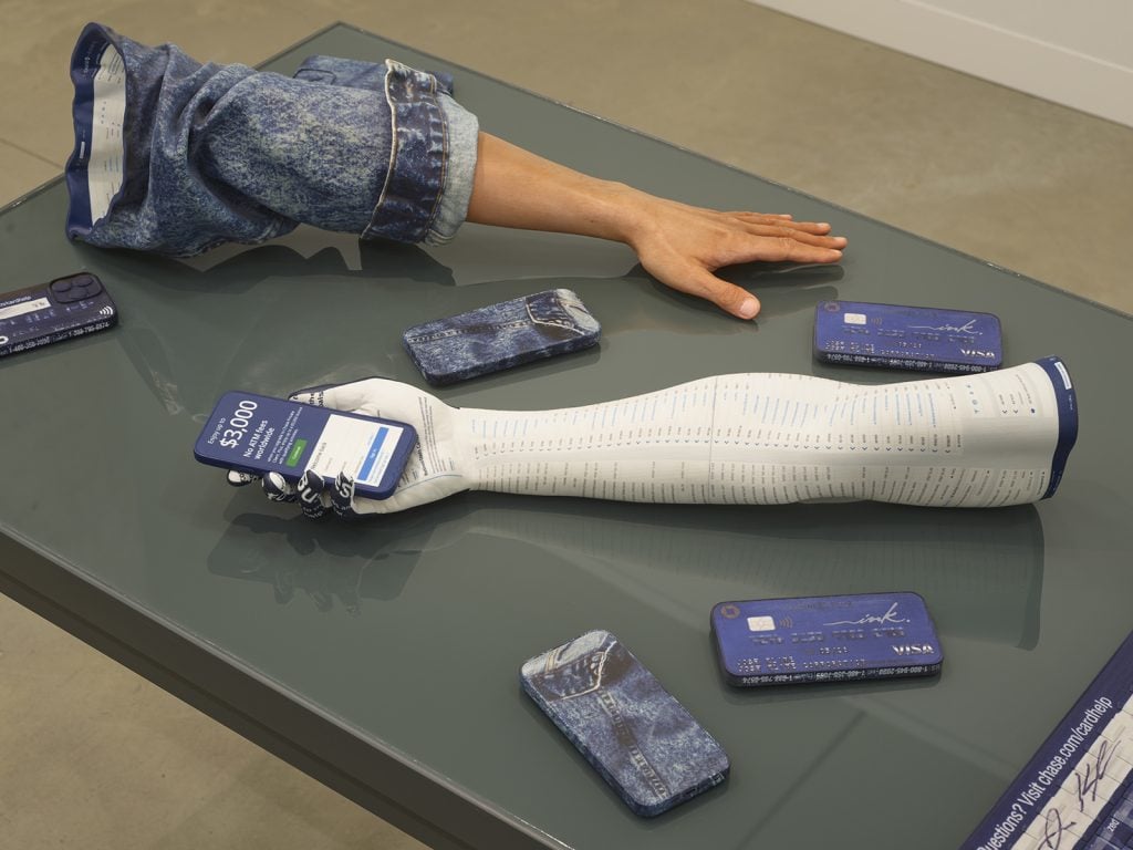 a disembodied arm in a denim jacket and the cast of a white arm are set on a table alongside five iphones