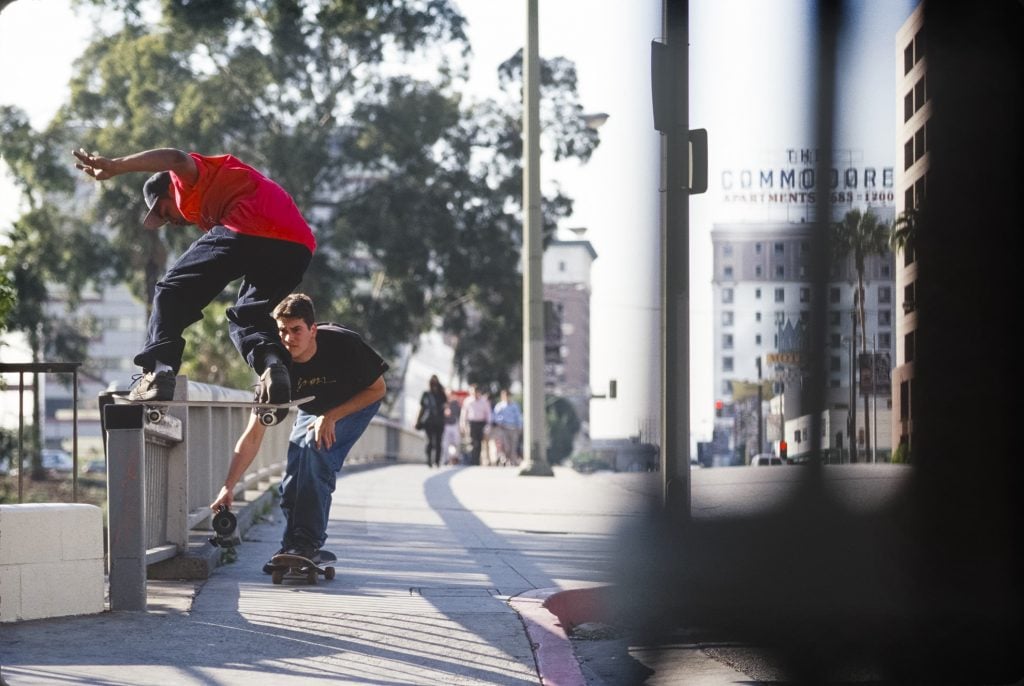 a street scene with a man in a red shirt on a skateboard and another filming him