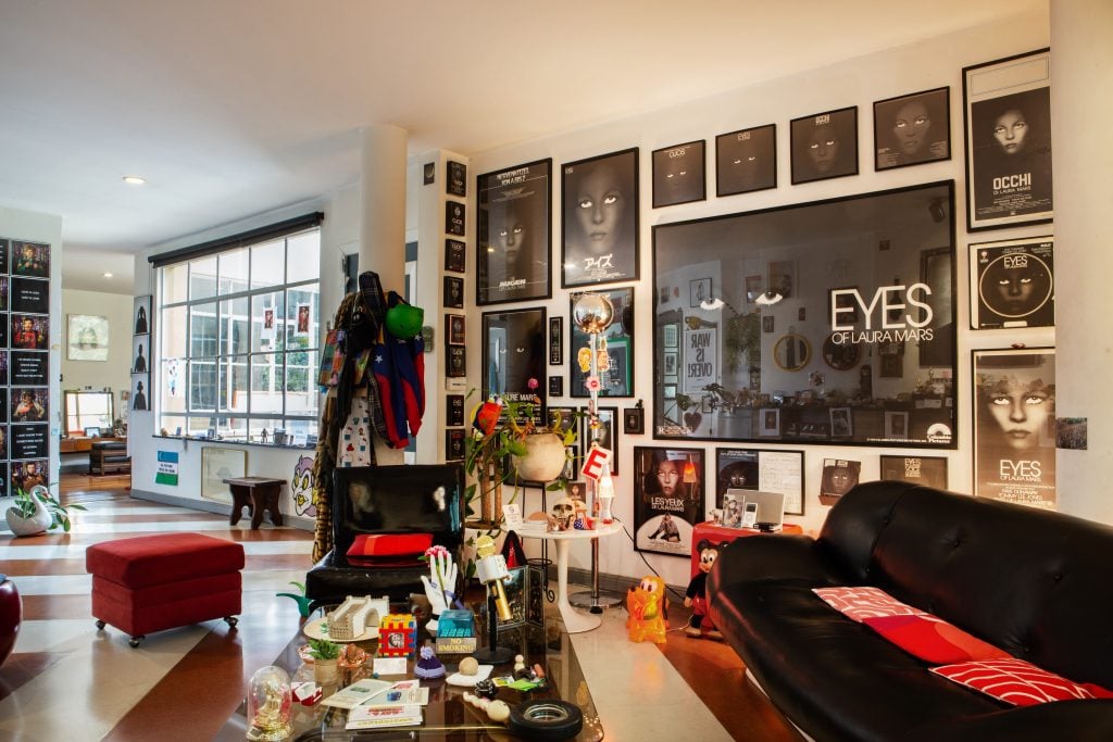 an apartment interior has a wall covered in ineternaiotnal posters of the 70s film 'the eyes of Laura mars' and retro modern furniture 