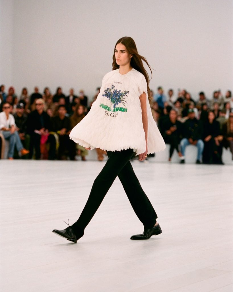 a model walking down the runway is wearing black trousers and a white flared top depicting van gogh's irises as attendees look on from behind her