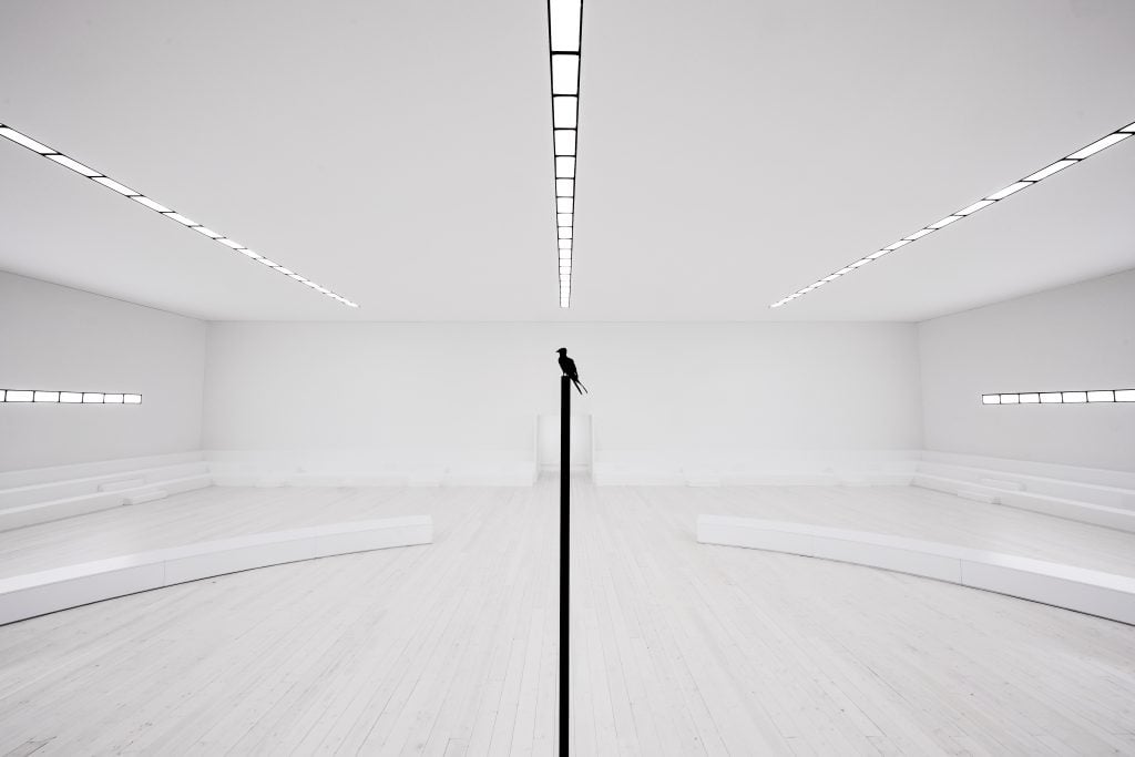 a delciate scultupre of a bronze bird perched on a pole stands in the center of an entirely white, plain room, surrounded by empty white benches.