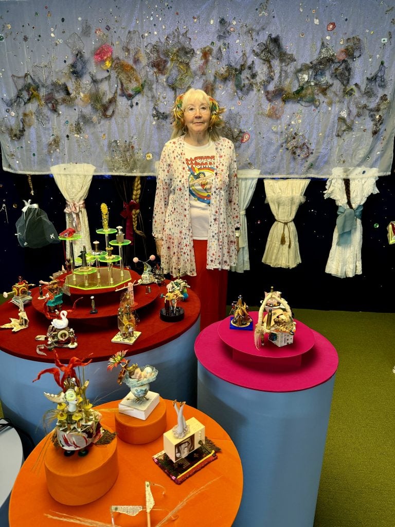 LaThoriel Badenhausen with her work at Spring Break Art Show New York 2024. An old white lady stands in front of a wall of textile work, behind three pedestals covered in tiny assemblage sculpture made from found objects. 