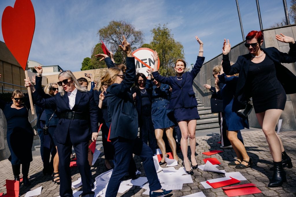 a group fo women wearing navy blue suits jump up and celebrate dancing on sheets of paper that have been thrown to the floor, a sign signalling 'no men' can be seen behind