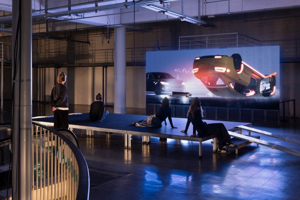 a large exhibition space awash with a glow of purple light and with a platform visitors can sit on, they are four people sitting on it, they are looking at a screen on which a car is seen turned upside down, it is clearly a film that they are watching