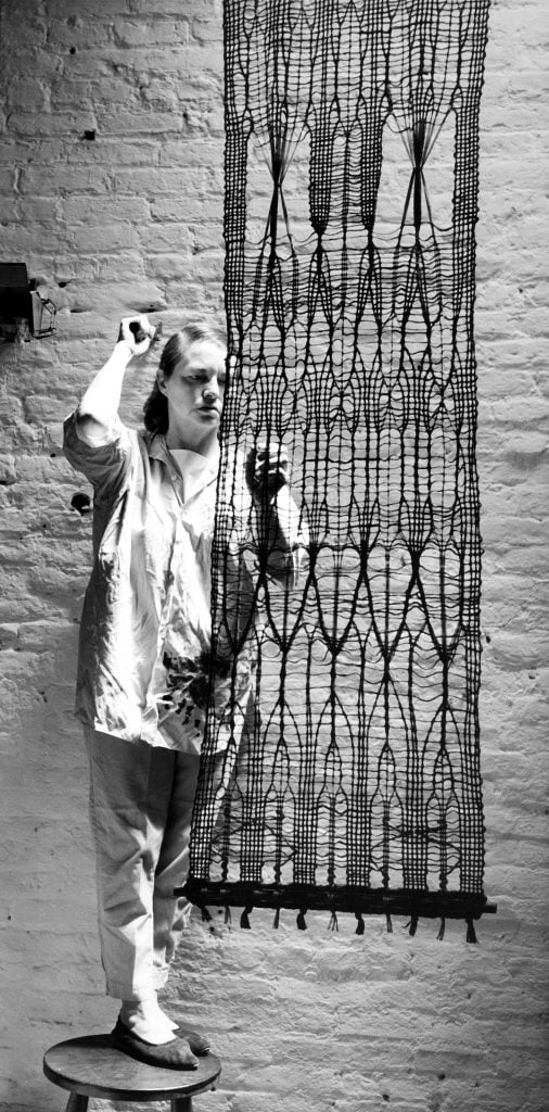 a black-and-white photograph of a woman in a buttondown shirt adjusting a fiber artwork.