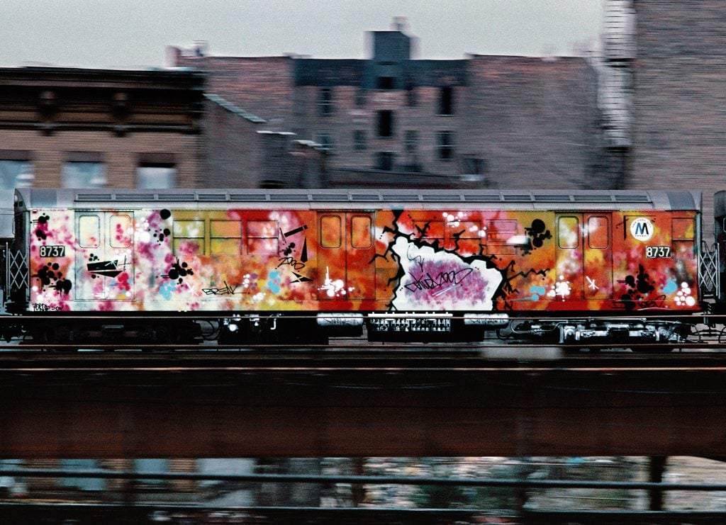 Martha Cooper Photograph of FUTURA 2000's Break Train (1980). A brightly painted subway train car, covered in abstract spray painted design, speeds down an elevated train track in the Bronx, the buildings behind slightly out of focus.