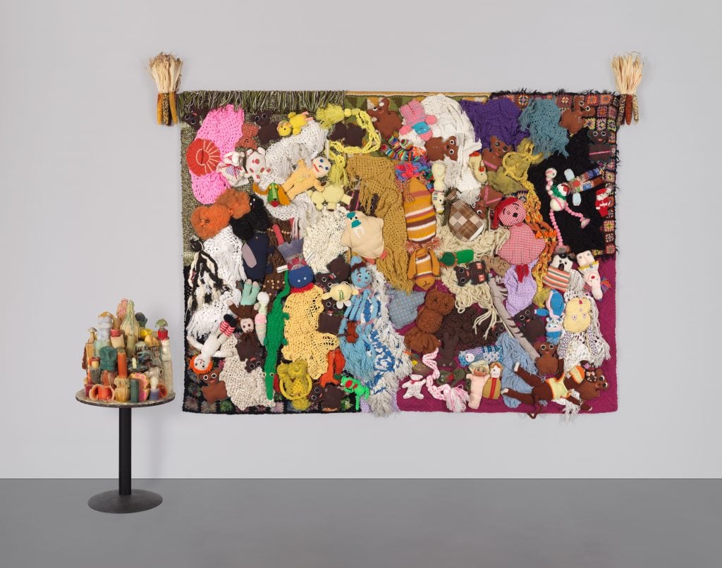 This image features a large, colorful tapestry made up of a dense collection of handmade and crocheted dolls, plush toys, and fabric scraps arranged in a chaotic, patchwork style. The tapestry is mounted on a wall and stretches horizontally across the frame, with various textures and colors, including pink, purple, yellow, and brown. The toys and dolls are intricately layered, creating a sense of abundance and overwhelming detail. To the left of the tapestry, there is a small round pedestal table displaying a variety of wax figures, arranged in a tower-like structure, adding to the eclectic and tactile quality of the artwork. The overall scene evokes themes of nostalgia, handmade craftsmanship, and the accumulation of personal memories.