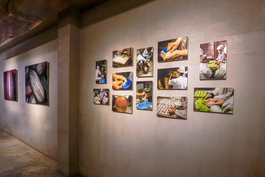 An installation photo of "Nathan Myhrvold: Intention and Detail" at the Gallery by Odo, New York. Photos are seen on a gray gallery wall. On the left, are two large-format photographs of Japanese cooking ingredients taken using a microscope. On the right, a salon-style hang features smaller photos of people making mame daifuku, a traditional Japanese dessert of mochi-wrapped red bean paste, in Tokyo. 